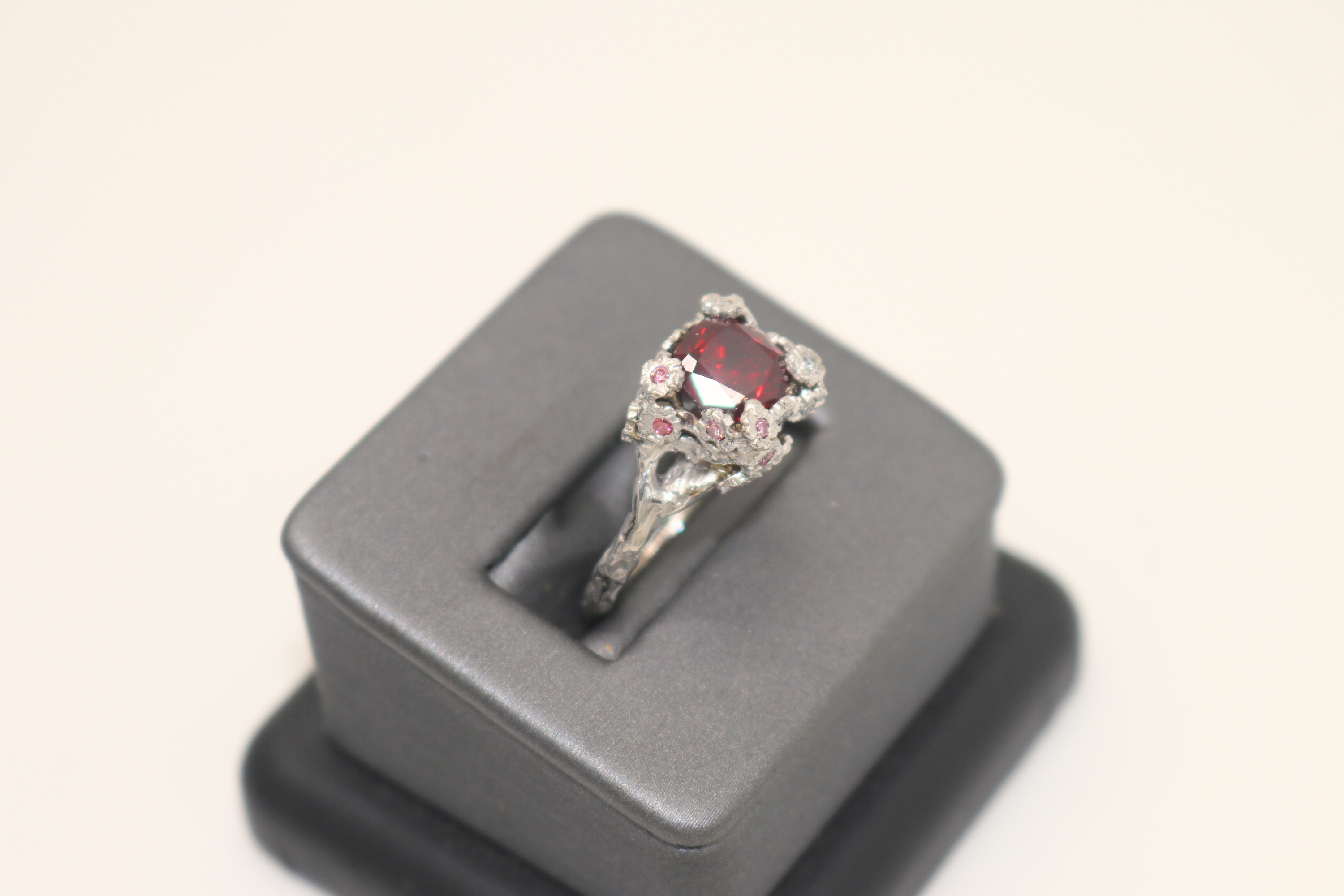 Foto de un anillo de compromiso con piedra central de granate rojo y diamantes rosas y blancos engastados en flores de oro blanco con una banda personalizada hecha para parecerse a una rama