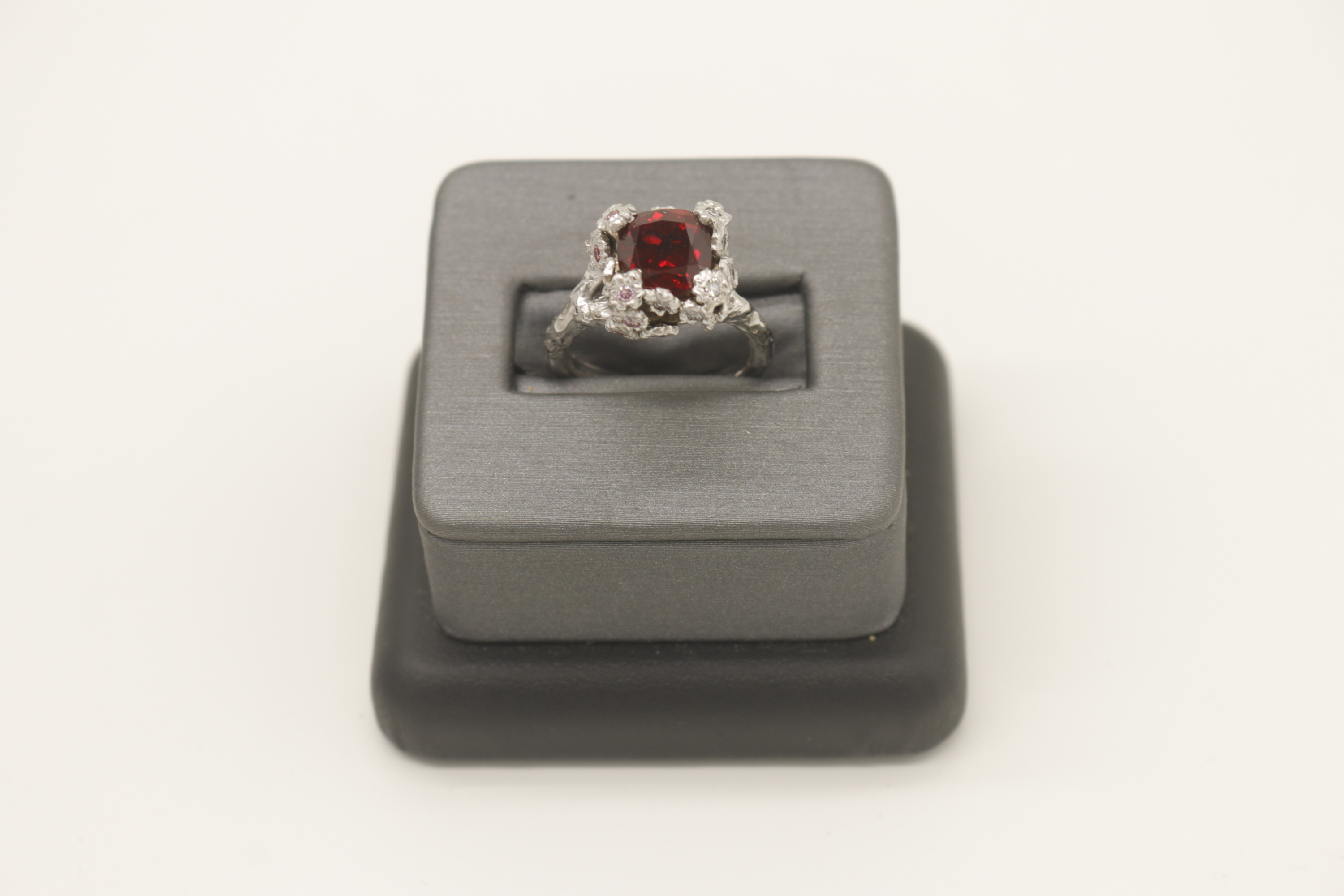 Foto de un anillo de compromiso con piedra central de granate rojo y diamantes rosas y blancos engastados en flores de oro blanco con una banda personalizada hecha para parecerse a una rama